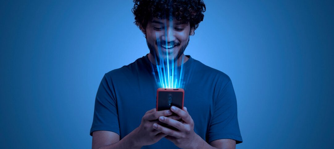 Homem sorrindo enquanto usa o celular, que emite raios azulados de uma interação Phygital.