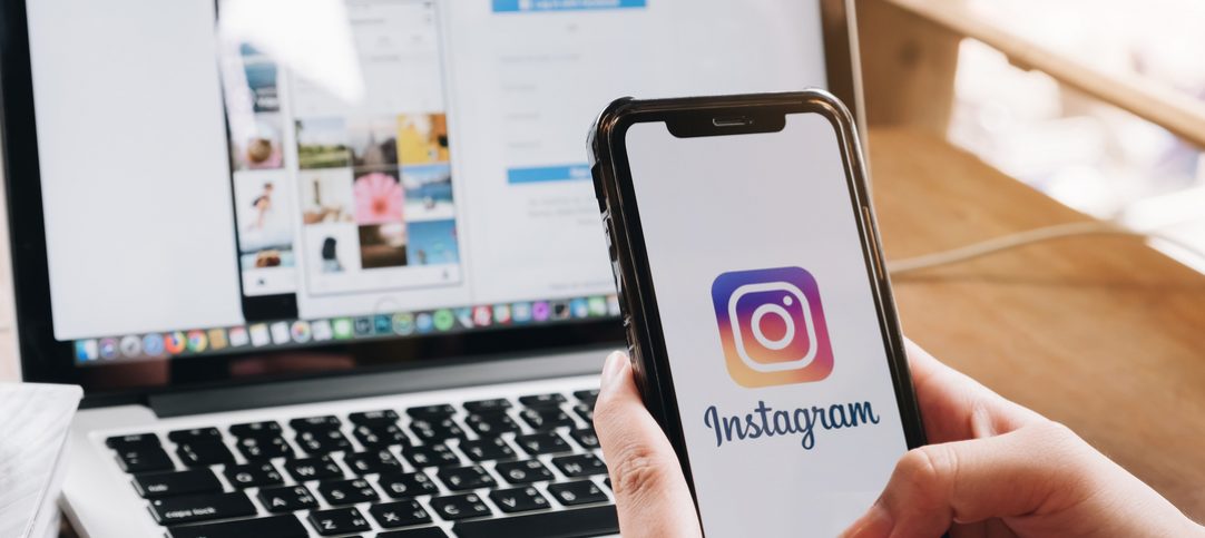Uma mulher segura um smartphone em frente a tela de um computador com o aplicativo do instagram aberto, prestes a fazer destaques no instagram.