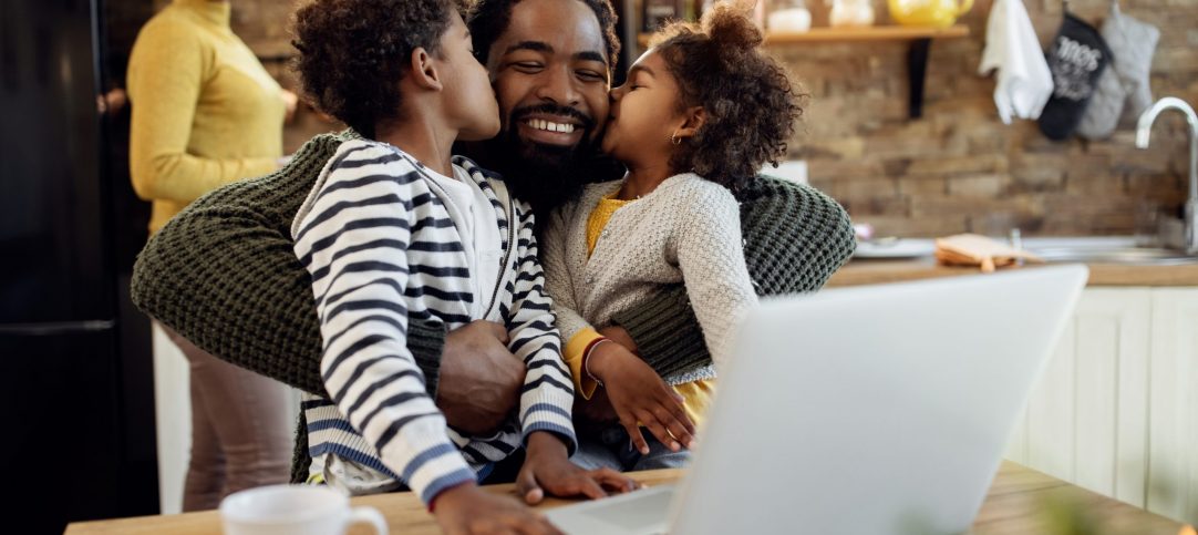 Happy black man being kissed by his children while using laptop at home. Mother is in the background.