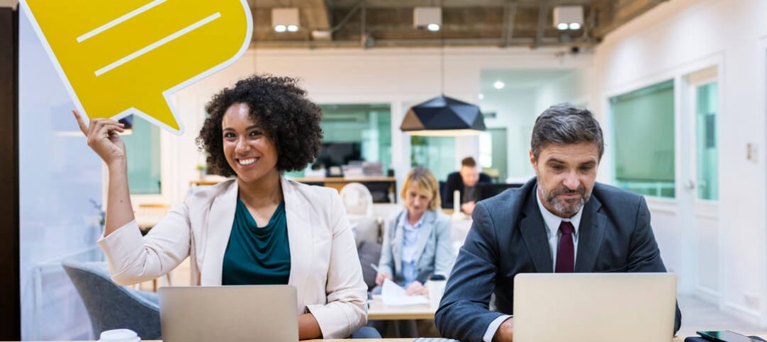 Homem e mulher sentados trabalhando em um computador. A mulher sorrindo com um balão de diálogo na mão, representando o marketing conversacional.